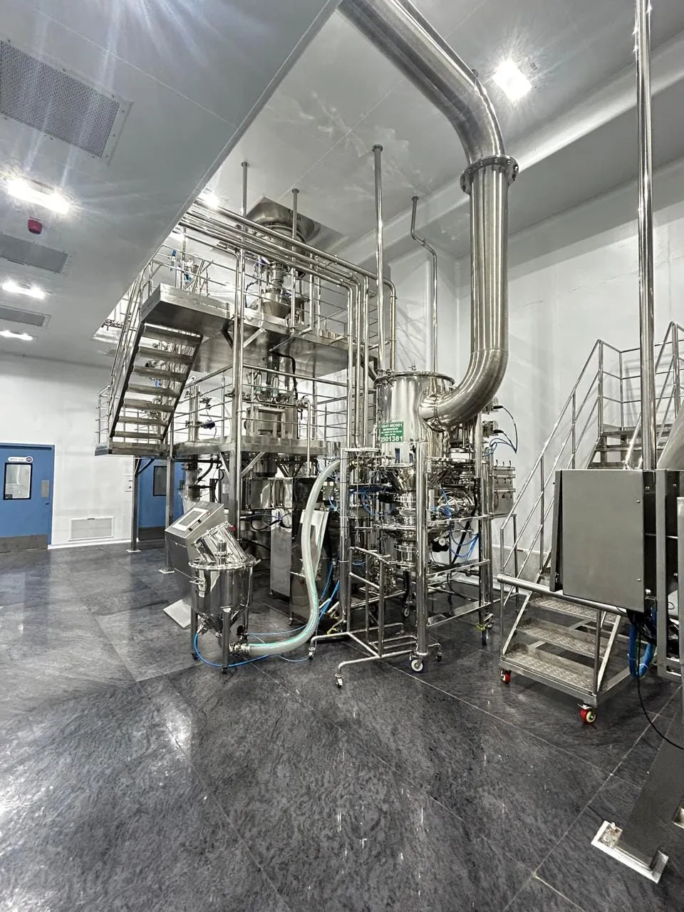 Glass-lined reactor train with a mezzanine access stair, seen across the tiled floor of a cGMP suite